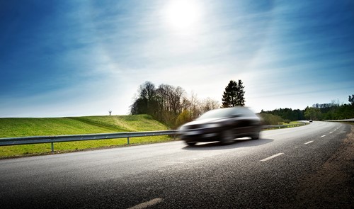 A car driving on a road with green fields and blue sky with a bright sun.