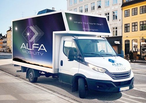 Vores designede Alfa-lastbil med Alfa Movings logo, som bruges til at transportere bohave, som befinder sig på en vej med huse i baggrunden.