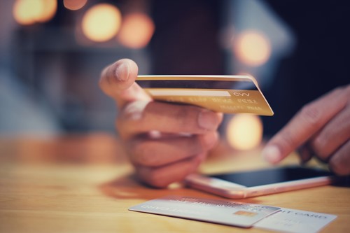 Et nærbillede af en hånd ved et skrivebord, der holder et kreditkort med et andet kort på skrivebordet sammen med en mobiltelefon.