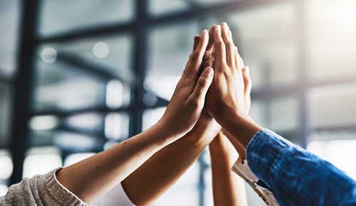 Five people joining hands in the air in an office setting.