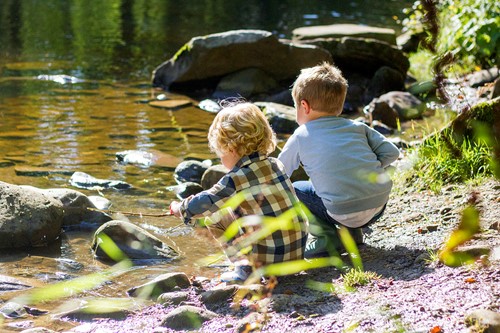 Two young boys sitting in the sand by the waterfront with rocks and greenery.