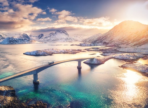 Vacker vy över ett landskap i Norge av en bro över havet samt snöiga berg i soldnedgång.