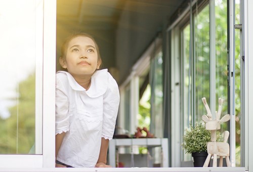 A young Asian girl looks out the window of her house at nature with a bright light in the sky.