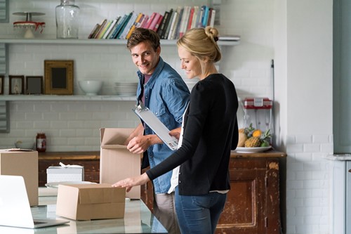 A smiling couple unpacking books standing by a book shelf in their apartment.