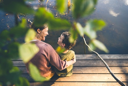 A laughing mother and son are sitting outside on a pier by the water with greenery in the foreground.