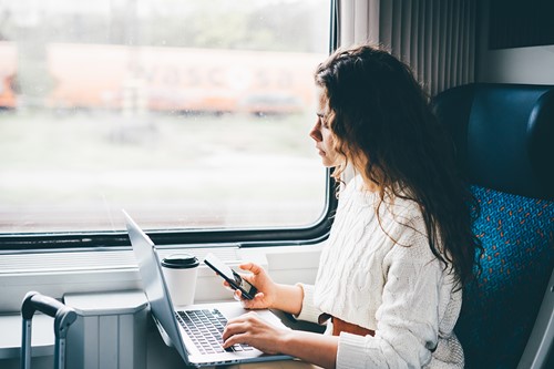 A woman traveling by train sitting by her computer holding a mobile phone.