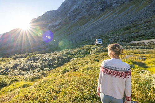 Nainen norjalaisessa villapaidassa seisoo vuoren juurella ja katselee aurinkoa kohti tielle tulevan kuorma-auton kanssa.