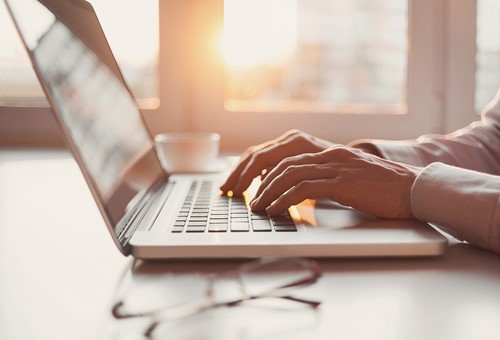 A close up of a person's hands typing on a computer at a desk with a pair of glasses laying on the desk.