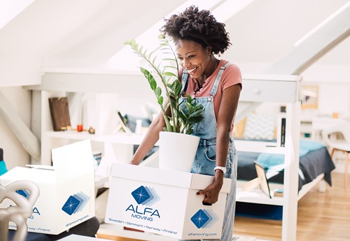 An african american woman carrying an white Alfa moving box with blue Alfa logo into her apartment.