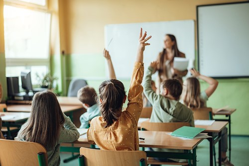 Et lyst klasserom med en lærer og elever som sitter ved pultene sine og rekker opp hendene.