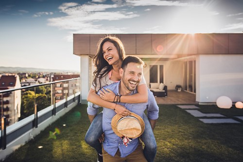 A husband carrying his spouse on his back outside their home with blue skies and a bright sun.