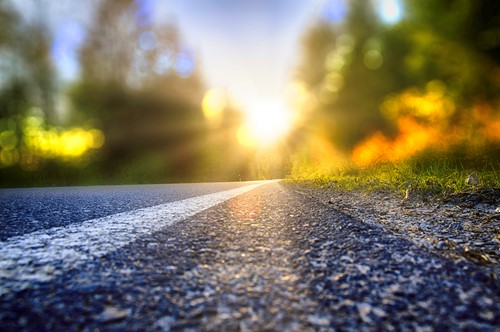 A close-up of a road with a view towards greenery and a bright sun.